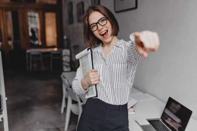 Positive business woman smiles and points finger at camera. Woman in pants and blouse poses with documents in office background.